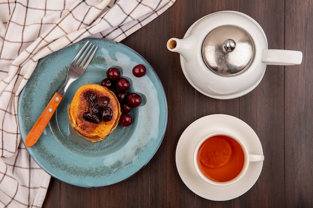 Draufsicht des Frühstückssatzes mit Pfannkuchen und Kirschen und Gabel in Platte auf kariertem Stoff und Tasse Tee mit Teekanne auf hölzernem Hintergrund