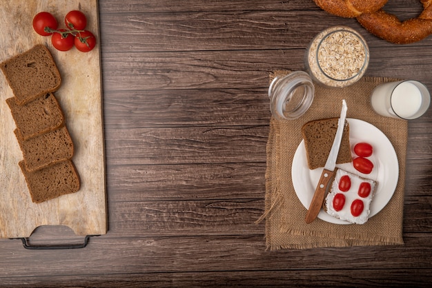 Draufsicht des Frühstückssatzes mit Roggenbrotscheiben, die mit Hüttenkäse und Tomaten mit Milch und Haferflocken auf hölzernem Hintergrund mit Kopienraum verschmiert wurden