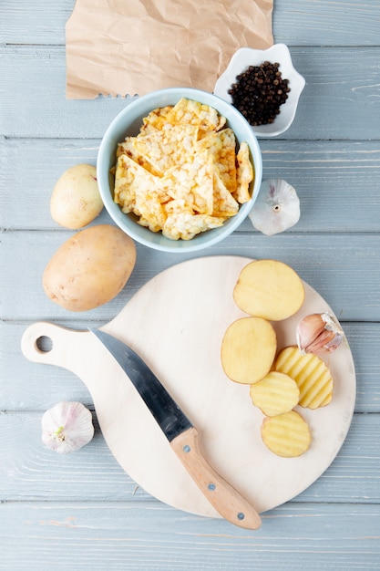 Draufsicht des gemüses als geschnittener kartoffelknoblauch mit messer auf schneidebrett und chips mit schwarzem pfeffer auf hölzernem hintergrund