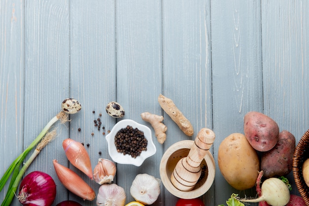 Draufsicht des Gemüses als Zwiebelkartoffelei-Knoblauch-Ingwer mit schwarzem Pfeffer und Knoblauchbrecher auf hölzernem Hintergrund mit Kopienraum