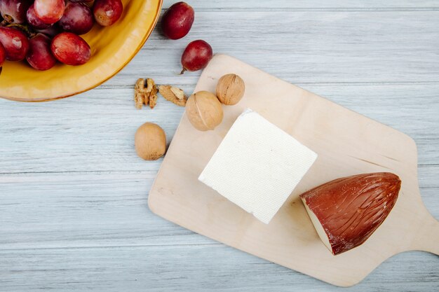 Draufsicht des geräucherten Käses und des Feta-Käses auf einem hölzernen Schneidebrett mit Walnüssen und süßen Trauben auf rustikalem Tisch