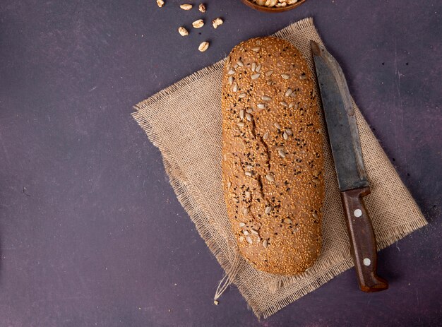 Draufsicht des gesäten Baguettes und des Messers auf Sackleinen auf der rechten Seite und kastanienbraunem Hintergrund mit Kopienraum