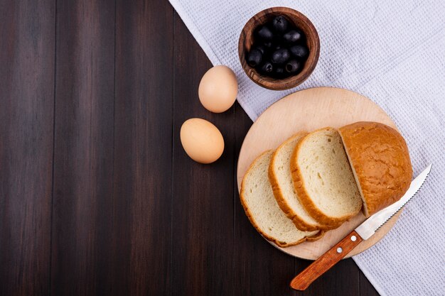 Draufsicht des geschnittenen Brotes und des Messers auf Schneidebrett mit Eiern und Schüssel der schwarzen Olive auf Stoff und Holzoberfläche