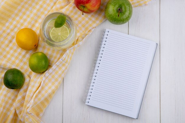 Draufsicht des Glases Wasser mit Limette und Zitrone auf einem gelben karierten Handtuch mit einem Notizbuch auf einer weißen Oberfläche
