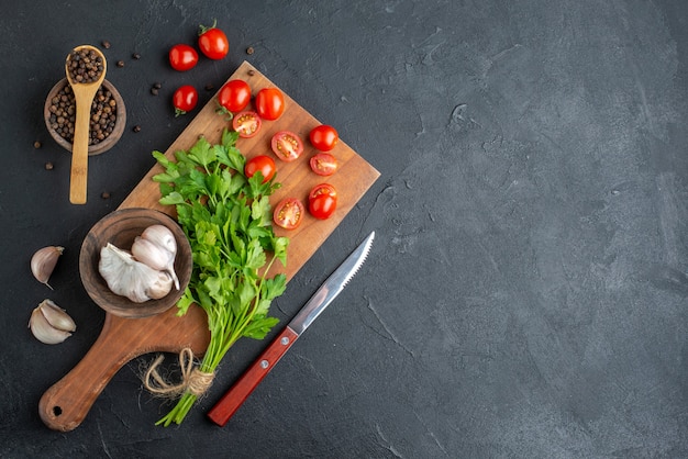 Draufsicht des grünen Bündels frischer ganz geschnittener Tomaten Knoblauch auf Holz Schneidebrett Messer Pfeffer auf schwarzer notleidender Oberfläche