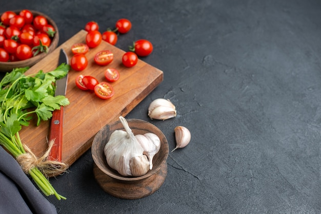 Draufsicht des grünen Bündels frischer Tomaten auf Holzschneidebrett und in Schüssel Knoblauch auf der rechten Seite auf schwarzer notleidender Oberfläche