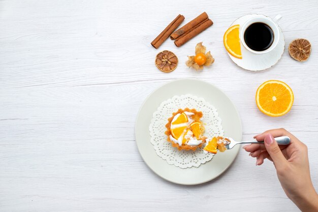 Draufsicht des kleinen Kuchens mit Sahne und geschnittenen Orangen, die von Frau zusammen mit Kaffee und Zimt auf hellem Schreibtisch, Obstkuchen süßer Zucker gegessen werden