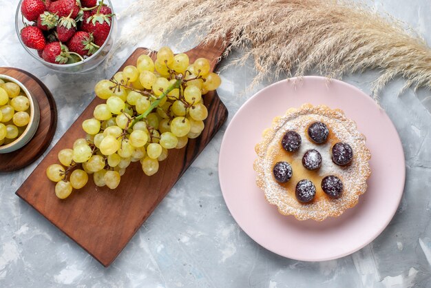 Draufsicht des kleinen Kuchens mit Zuckerpulverfrüchten zusammen mit frischen roten Erdbeeren und Trauben auf leichtem süßem Tee des Obstkuchenbackens