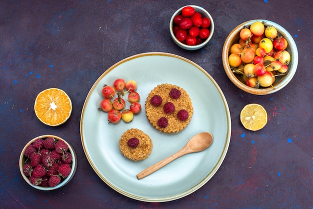 Draufsicht des kleinen runden Kuchens mit frischen Himbeeren innerhalb Platte mit frischen Früchten auf dunkler Oberfläche