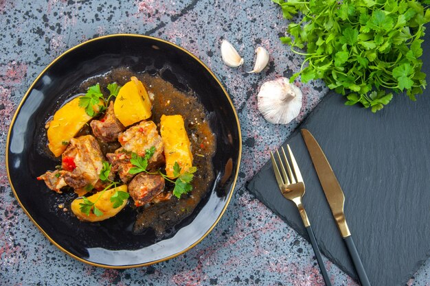 Draufsicht des köstlichen Abendessens mit Fleischkartoffeln serviert mit Grün in einem schwarzen Teller und Besteck auf Schneidebrett Knoblauch auf Mischfarben Hintergrund