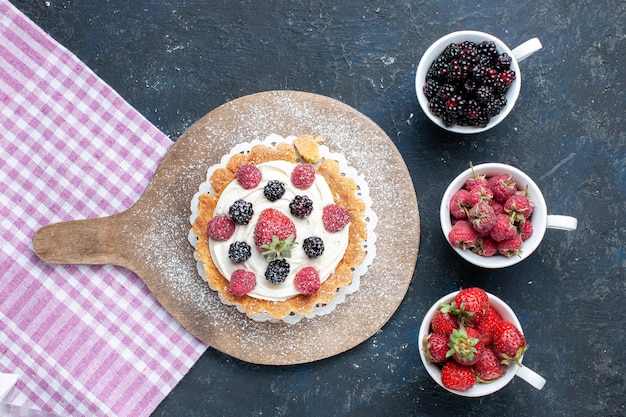 Kostenloses Foto draufsicht des köstlichen kleinen kuchens mit zuckerpulvercreme und -beeren zusammen mit tassen beeren auf dunklem schreibtisch, beerenfruchtkuchen-keks