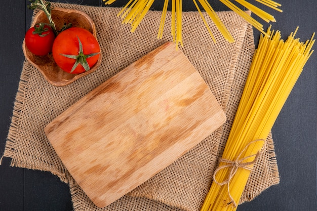 Draufsicht des Küchenbretts mit rohen Spaghetti und Tomaten auf einer beigen Serviette auf einer schwarzen Oberfläche