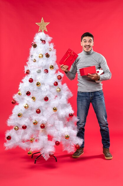 Draufsicht des lächelnden gutaussehenden Erwachsenen in einer grauen Bluse, die nahe dem verzierten weißen Weihnachtsbaum steht