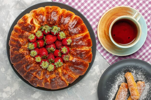 Draufsicht des leckeren Erdbeerkuchens mit frischen roten Erdbeeren und einer Tasse Tee auf weißem Schreibtisch