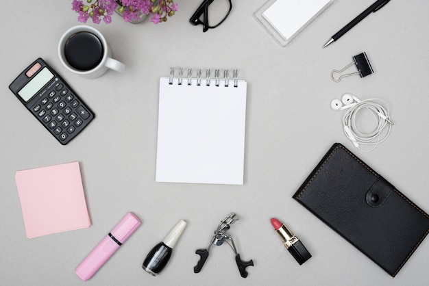 Draufsicht des leeren Notizblockes umgeben durch Kaffeetasse; Taschenrechner; Make-up-Objekte und Kopfhörer auf grauem Hintergrund