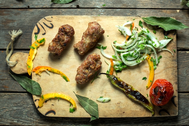 Draufsicht des Lula Kebab mit Zwiebelkräutern und gegrilltem Gemüse auf einem Holzbrett