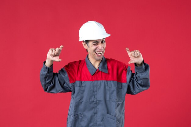 Draufsicht des lustigen jungen Baumeisters in Uniform mit Schutzhelm, der etwas auf isolierter roter Wand erklärt