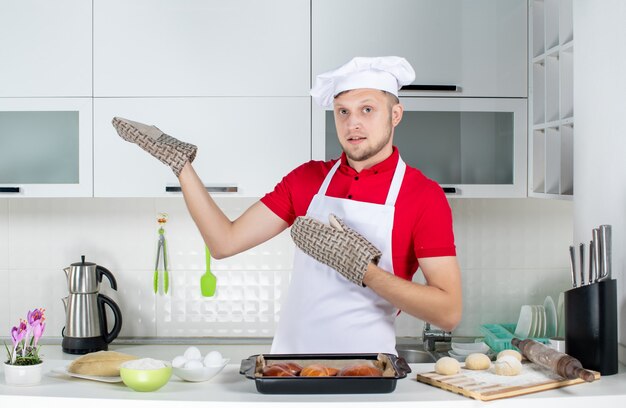 Draufsicht des männlichen Kochs, der einen Halter trägt, der hinter dem Tisch mit Gebäck-Eierreibe steht und etwas auf der rechten Seite in der weißen Küche zeigt