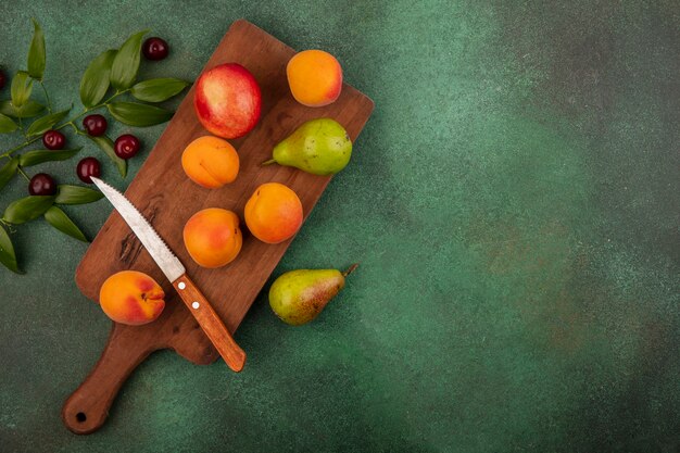 Draufsicht des Musters von Früchten als Aprikosenpfirsich und Birne mit Messer auf Schneidebrett und Kirschen mit Blättern auf grünem Hintergrund mit Kopienraum