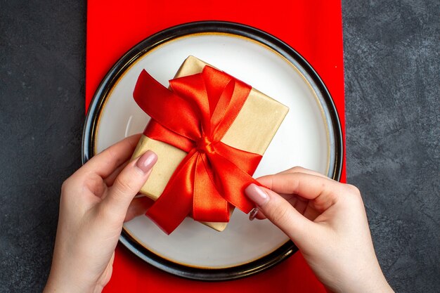 Draufsicht des nationalen Weihnachtsmahlzeithintergrundes mit Hand, die leere Teller mit bogenförmigem rotem Band auf einer roten Serviette auf schwarzem Tisch hält