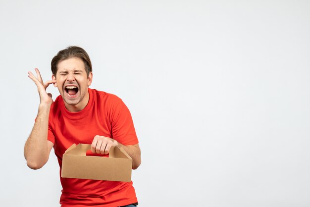 Draufsicht des nervösen und emotionalen jungen Mannes in der roten Bluse, die Kasten hält, der einen seiner Ohren auf weißem Hintergrund schließt