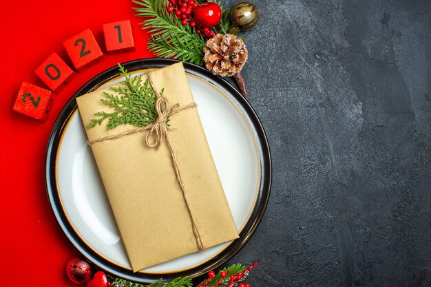 Draufsicht des Neujahrshintergrunds mit Geschenk auf Tellerdekorationszubehör Tannenzweigen und Zahlen auf einer roten Serviette auf der rechten Seite auf einem schwarzen Tisch