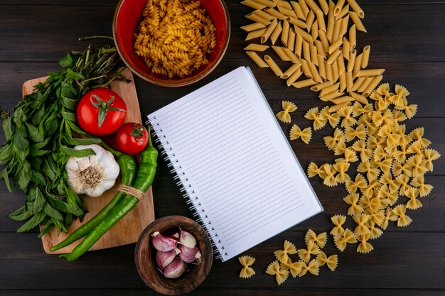 Draufsicht des Notizbuchs mit rohen Nudeltomaten Knoblauch mit Chili-Pfeffer und Minze auf einem Schneidebrett auf einer Holzoberfläche