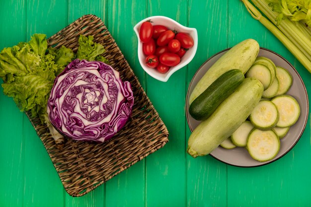 Draufsicht des Purpurkohls auf einem Weidentablett mit Salat mit Zucchini und Gurke auf einem Teller mit Pflaumentomaten auf einer Schüssel mit Sellerie lokalisiert auf einer grünen Holzwand