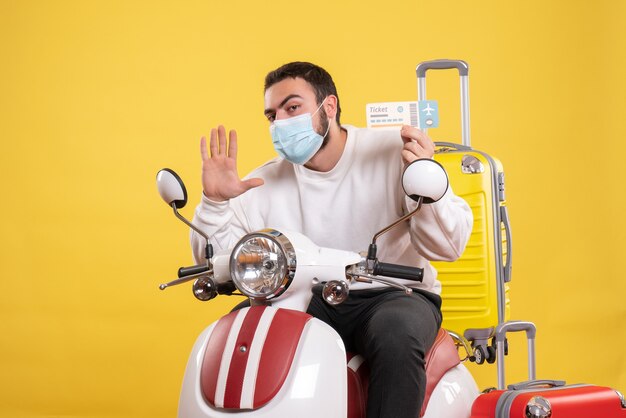 Draufsicht des Reisekonzepts mit einem jungen Mann in medizinischer Maske, der auf einem Motorrad mit gelbem Koffer darauf sitzt und ein Ticket mit fünf hält