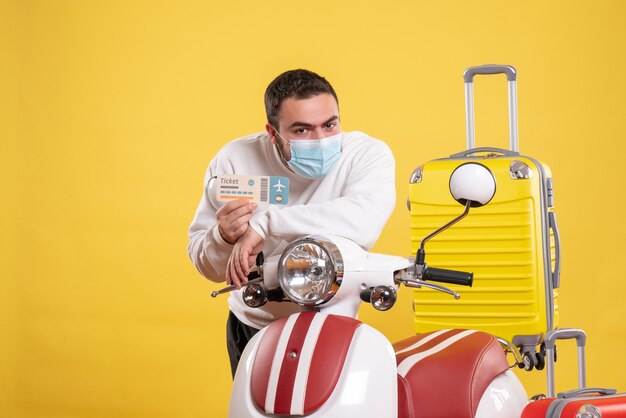 Draufsicht des Reisekonzepts mit fokussiertem Kerl in medizinischer Maske, der in der Nähe eines Motorrads mit gelbem Koffer steht und ein Ticket hält