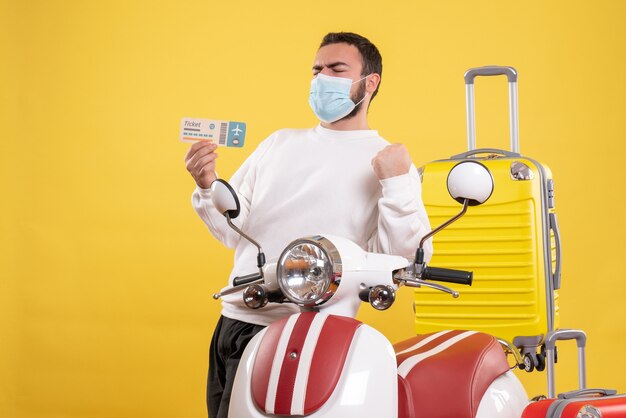 Draufsicht des Reisekonzepts mit stolzem Kerl in medizinischer Maske, der in der Nähe eines Motorrads mit gelbem Koffer steht und ein Ticket hält