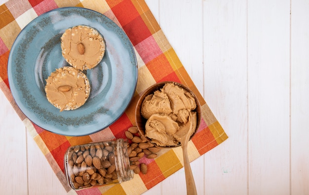 Draufsicht des Reisgetreidecrackers mit Erdnusspastenbutter auf blauer Keramikplattenmandel, die von einem Glas und einer Schüssel mit Erdnussbutter auf karierter Tischserviette auf weißem hölzernem Hintergrund wi verstreut wird