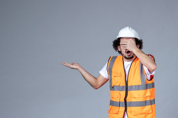 Draufsicht des schockierten männlichen Architekten in Warnweste mit Schutzhelm und nach oben zeigend auf der rechten Seite, der sein Auge auf der grauen Wellenwand schließt