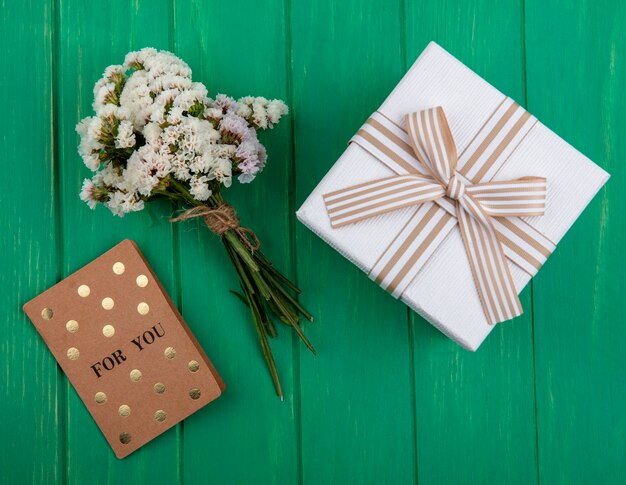 Draufsicht des Straußes der weißen Blumen mit einer braunen Karte und einem Geschenk in einer weißen Packung mit einer Schleife auf einer grünen Oberfläche