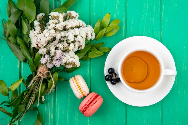 Draufsicht des Straußes der weißen Blumen mit einer Tasse Tee mit Macarons auf einer grünen Oberfläche