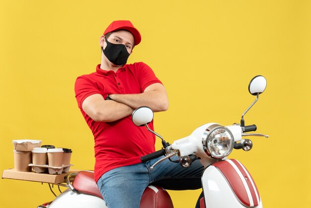 Draufsicht des verträumten jungen Erwachsenen, der rote Bluse und Huthandschuhe in der medizinischen Maske trägt, die Ordnung liefert, die auf Roller auf gelbem Hintergrund sitzt