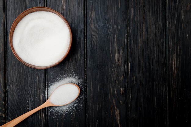 Draufsicht des weißen granulierten Zuckers in einer Holzschale und in einem Holzlöffel auf schwarzem rustikalem Hintergrund mit Kopienraum