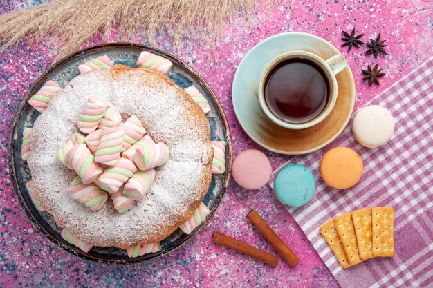 Draufsicht des Zuckerpulverkuchens mit Macarons und Tasse Tee auf rosa Oberfläche