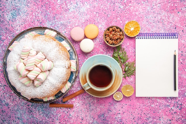 Draufsicht des Zuckerpulverkuchens mit Tasse Tee und französischen Macarons auf rosa Oberfläche