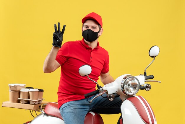 Draufsicht des zuversichtlichen jungen Erwachsenen, der rote Bluse und Huthandschuhe in der medizinischen Maske trägt, die Ordnung liefert, die auf Roller sitzt, der drei auf gelbem Hintergrund zeigt