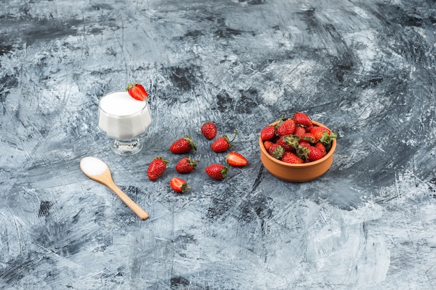 Draufsicht eine Glasschale Joghurt auf Weiden-Tischset mit Holzlöffel und Erdbeeren auf dunkelblauem Marmor und weißer Holzbrettoberfläche. vertikal