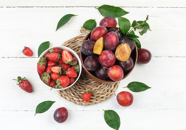 Draufsicht eine Schüssel Pflaumen und Erdbeeren auf Weiden Tischset auf weißem Holzbrett Tisch. horizontal