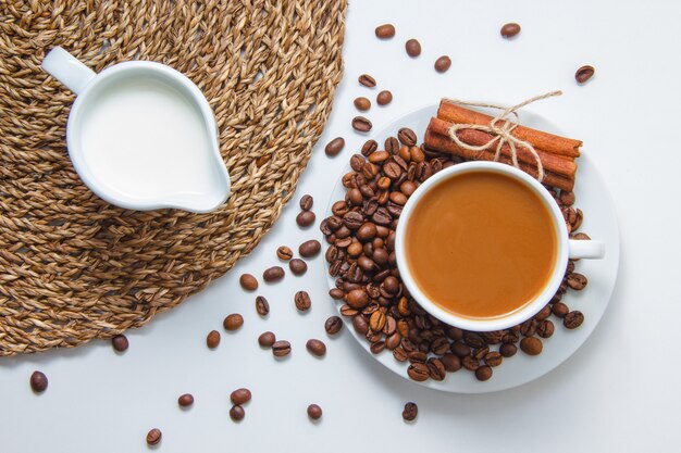 Draufsicht eine Tasse Kaffee mit Kaffeebohnen, Milch, trockenem Zimt auf Untersetzer und weißer Oberfläche. horizontal