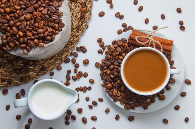 Draufsicht eine Tasse Kaffee mit Kaffeebohnen, Milch, trockenem Zimt auf Untersetzer und weißer Oberfläche. horizontal