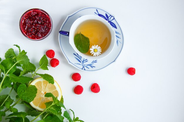 Draufsicht eine Tasse Kamillentee mit Zitrone, Minzblättern, Himbeeren, Marmelade in Untertasse auf Stoff