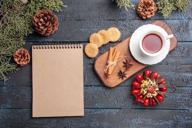 Draufsicht eine Tasse Tee Anissamen und Zimt auf hölzernem Servierteller Kekse Tannenzapfen Beerenkuchen und ein Notizbuch auf dunklem Holztisch