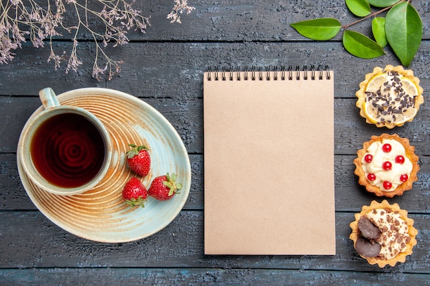 Draufsicht eine Tasse Tee und Erdbeeren auf Untertassen getrockneten Blumenzweigtörtchenblätter und ein Notizbuch auf dunklem Holztisch