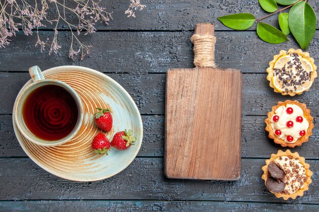 Draufsicht eine Tasse Tee und Erdbeeren auf Untertassen getrockneten Blumenzweigtörtchenblätter und ein Schneidebrett auf dunklem Holztisch