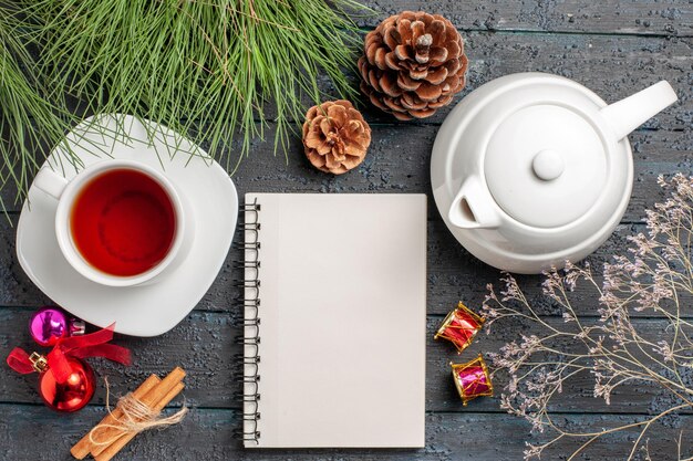 Draufsicht eine Tasse Tee weiße Tasse Tee Teekannenplätzchen neben den Zimtstangen weiße Notizbuch-Fichtenzweige mit Weihnachtsspielzeug und Zapfen auf dem Tisch