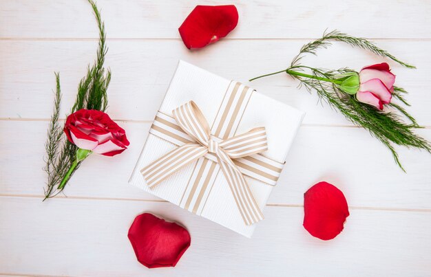 Draufsicht einer Geschenkbox gebunden mit Schleife und roten Farbrosen mit verstreuten Blütenblättern und Spargel auf weißem hölzernem Hintergrund